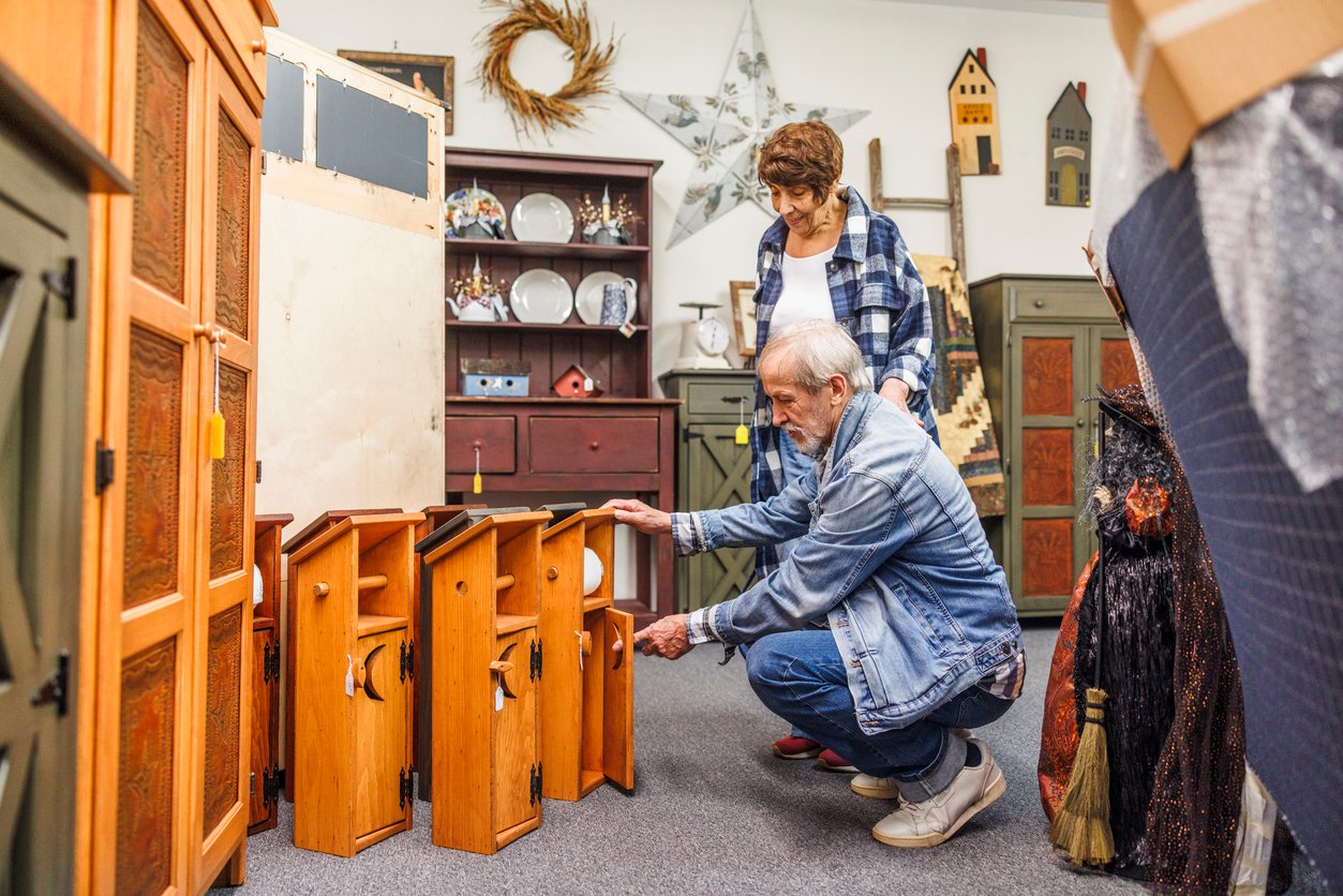 An older man kneels to examine wooden cabinets in a shop while an older woman stands behind him, both surrounded by rustic furniture and decorative items.