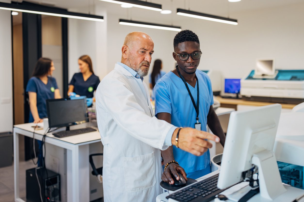 An older man in a white lab coat points at a computer screen while a younger man in blue scrubs watches. In the background, two women in scrubs talk near lab equipment in a modern laboratory setting.