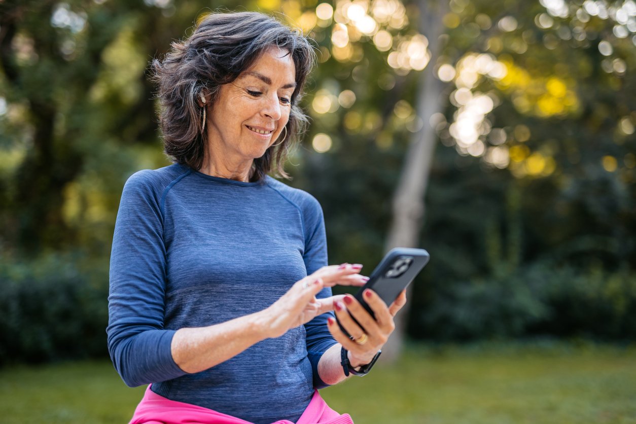 A woman with shoulder-length brown hair and wearing a blue long-sleeve shirt smiles while using her smartphone outdoors, surrounded by greenery and sunlight.