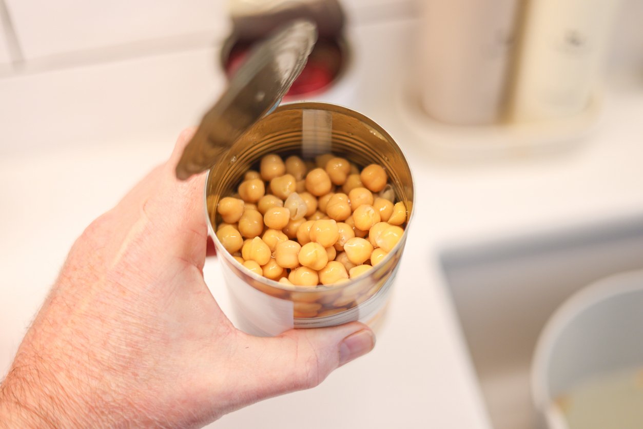 A hand holds an open can filled with chickpeas over a kitchen sink, with some kitchen items blurred in the background.