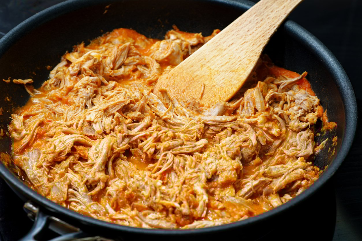 Shredded chicken cooked in a reddish-orange sauce, being stirred with a wooden spatula in a black skillet.