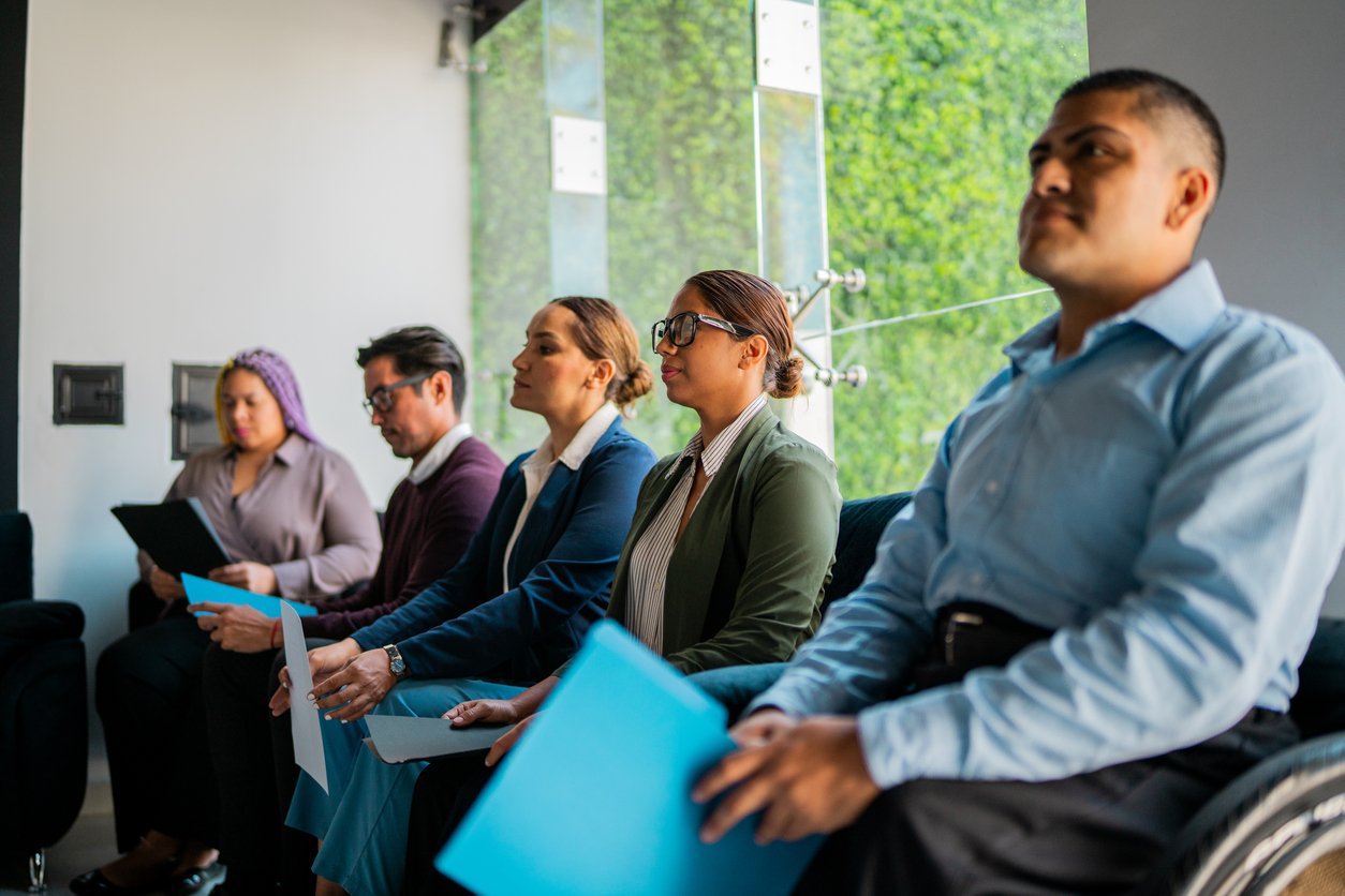 Five professionally dressed people sit in a row in an office, holding folders and laptops, appearing focused and waiting for an interview or meeting; one person uses a wheelchair.