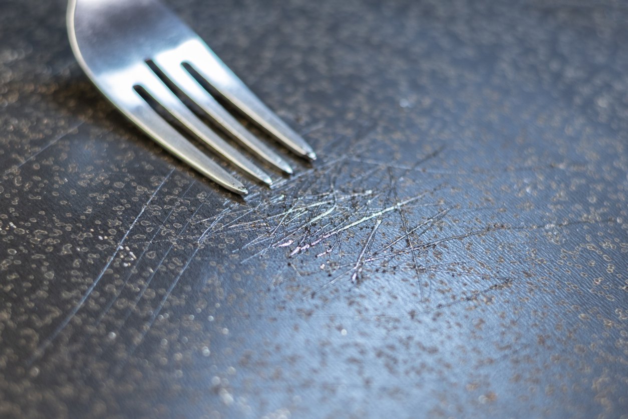 A close-up of a metal fork scraping the scratched surface of a dark non-stick pan, showing visible scrape marks and wear.