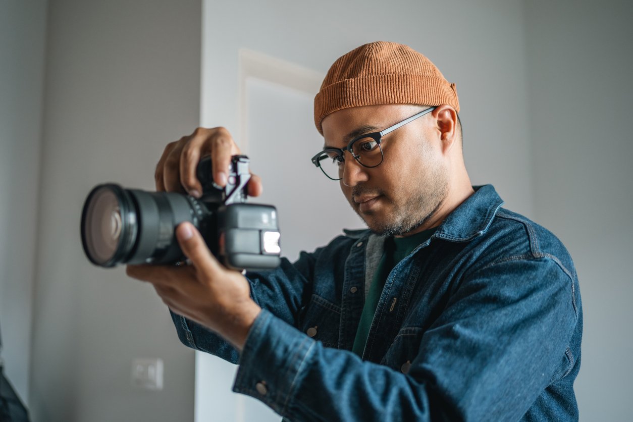 A person wearing glasses, a brown beanie, and a denim jacket holds a camera, looking intently at the viewfinder in a well-lit indoor space.