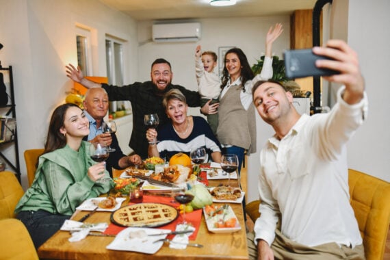 A group of six adults and one child smile and pose for a selfie around a dinner table filled with food, wine, and dessert, capturing the joy—and perhaps reflecting on the thanksgiving dinner cost—of a cozy, well-lit dining room.