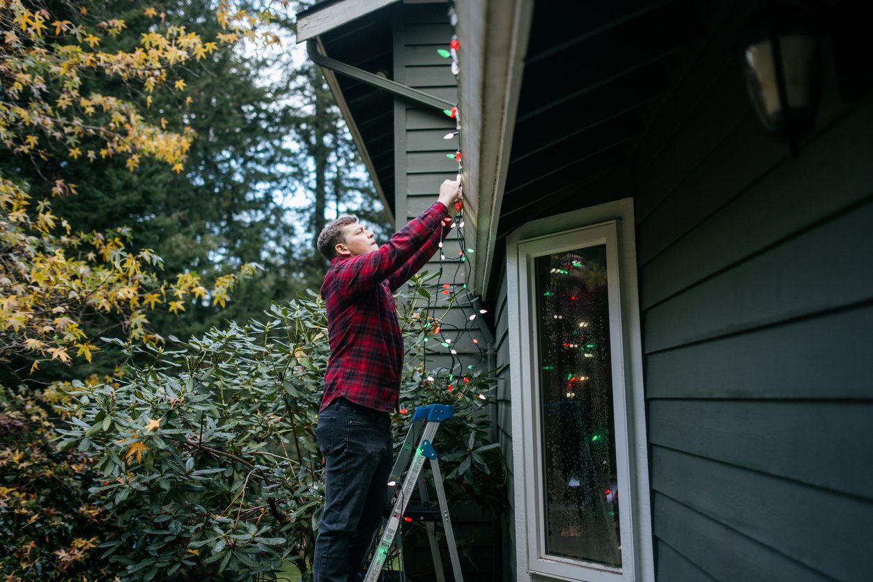 A person standing on a ladder hangs colorful string lights along the roof of a house, preparing holiday decorations outdoors near trees and bushes.