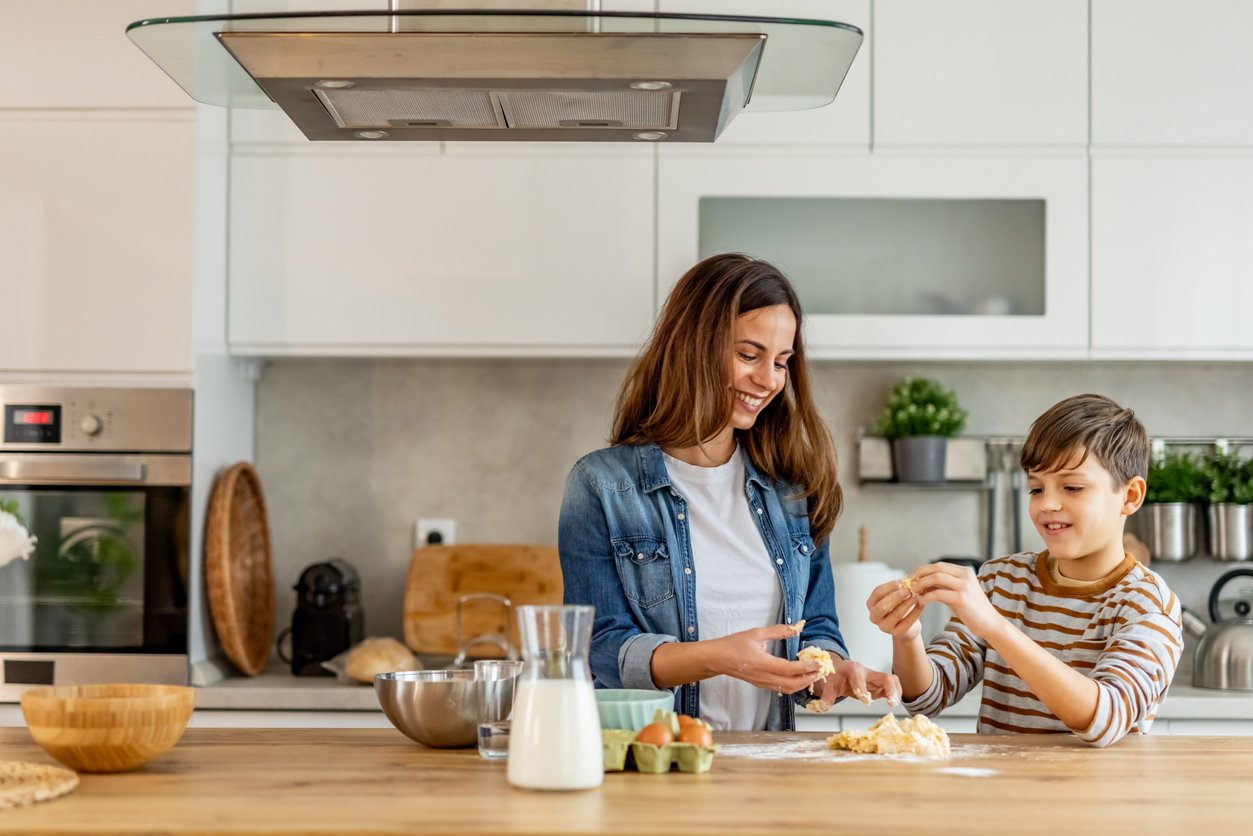 A woman and a young boy smile while making dough together in a bright, modern kitchen. Mixing bowls, eggs, and a glass of milk are on the wooden counter in front of them.