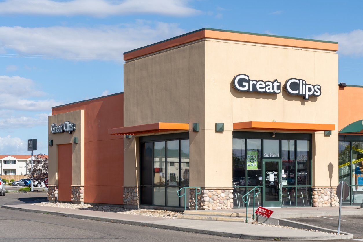 A Great Clips hair salon in a modern, tan-colored building with large windows and orange trim, located in a shopping plaza on a sunny day.
