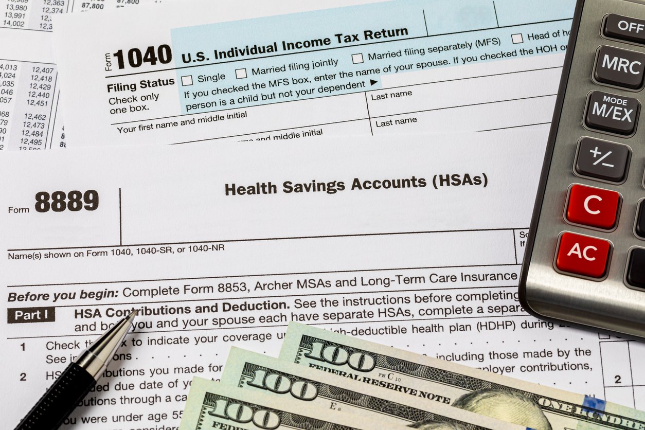 A U.S. 1040 tax form, Form 8889 for Health Savings Accounts, a calculator, a pen, and several $100 bills are arranged on a desk, representing tax preparation and financial planning.