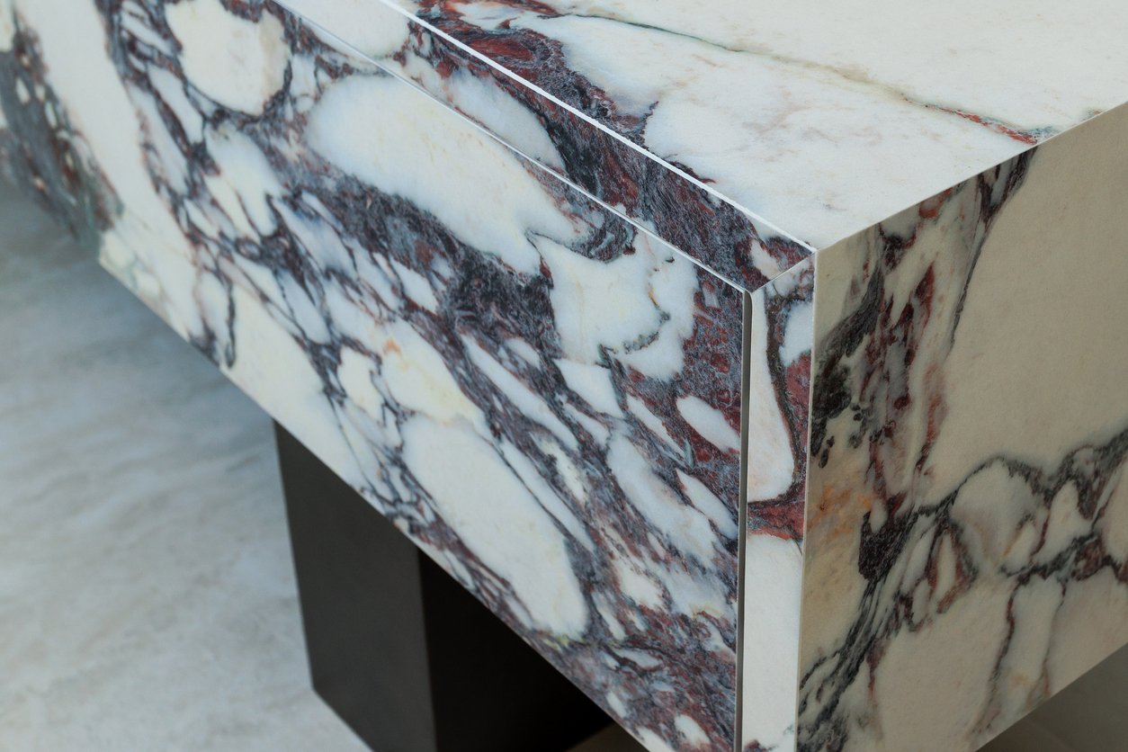 Close-up of a modern marble table with bold, intricate veining in black, white, and reddish tones. The table has sharp edges and is supported by dark rectangular legs on a light-colored floor.