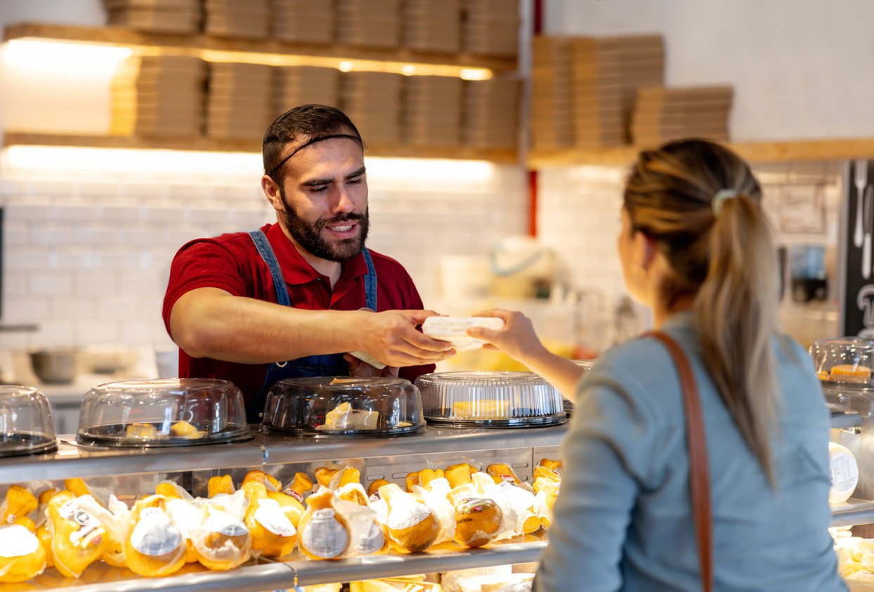 A bakery worker in a red shirt and apron hands a pastry to a customer across a glass display filled with baked goods. The customer is seen from behind, and shelves with boxes are visible in the background.