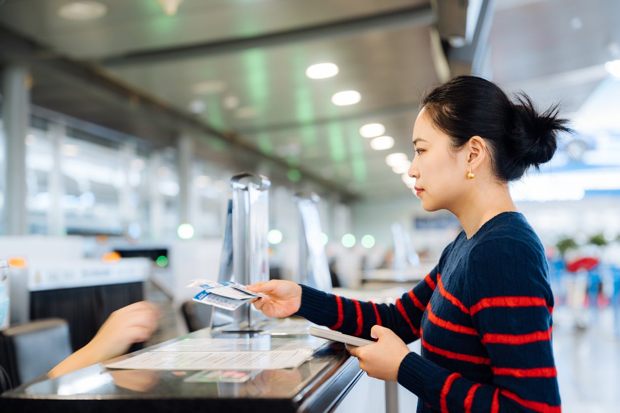 A woman in a blue and red striped sweater stands at an airport counter, handing over travel documents or tickets to an employee behind the desk. The setting is bright with a modern interior.