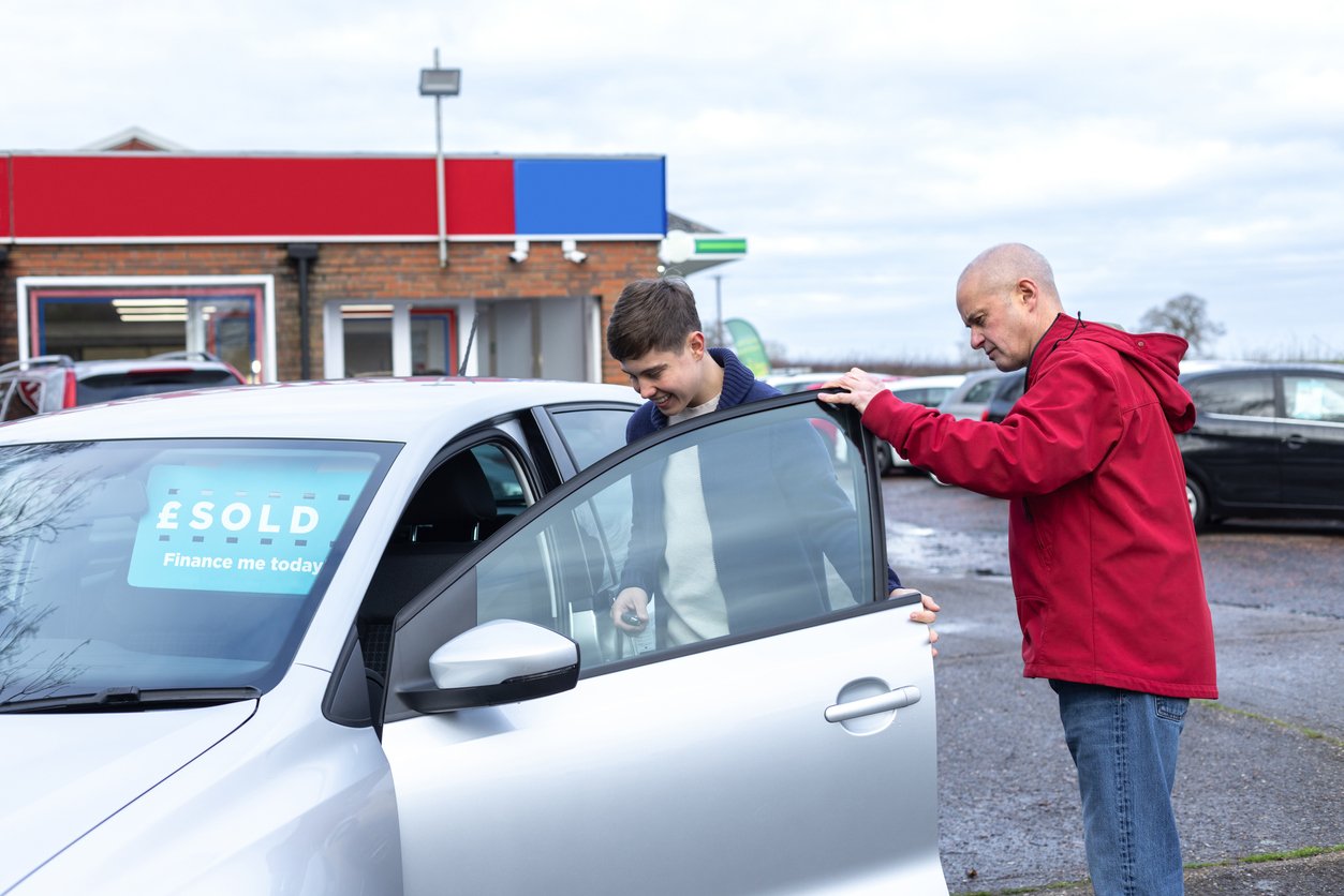 A young man opens the door of a silver car with a "SOLD" sign on the windshield, while an older man in a red jacket stands next to him outside a dealership.