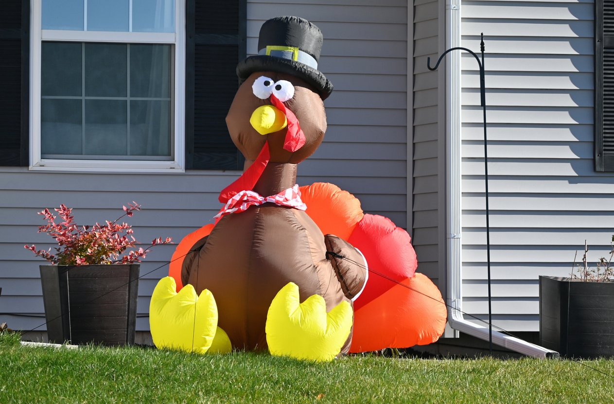 An inflatable turkey with a black hat and red scarf sits on a lawn in front of a house, hinting at Thanksgiving dinner cost history and adding festive cheer to the gray siding and potted plants.