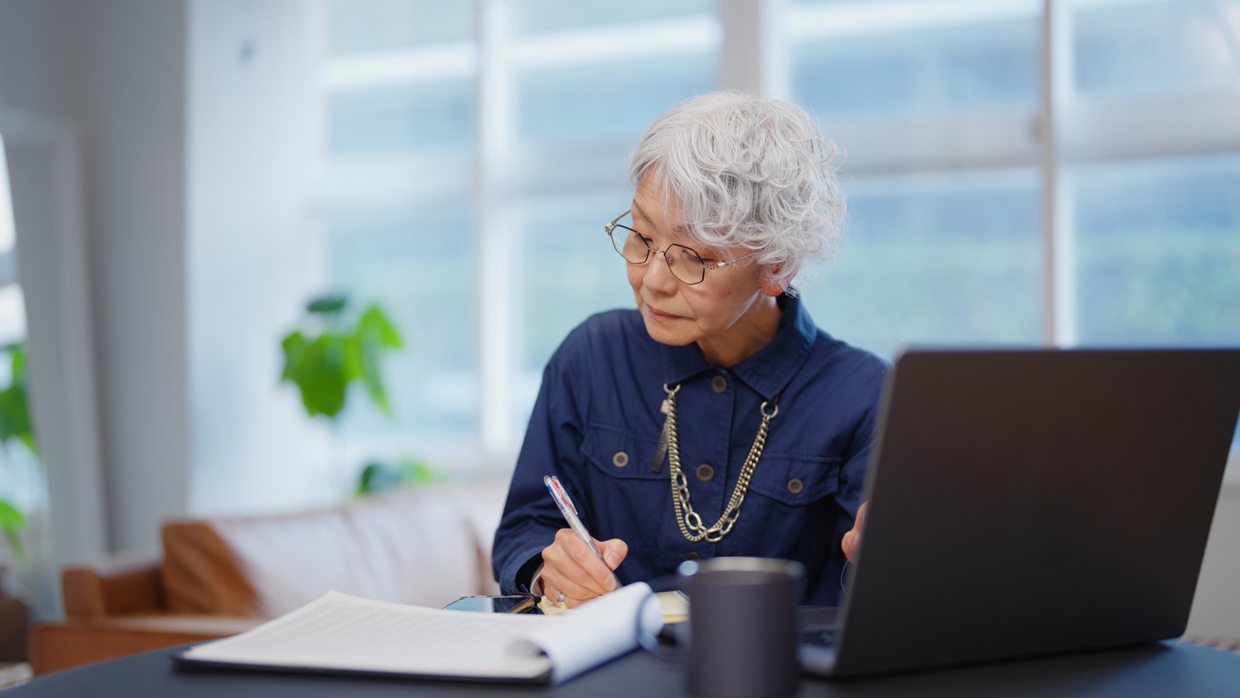 An older woman with short gray hair and glasses sits at a desk, writing in a notebook beside an open laptop. She is wearing a blue blouse and necklace, with a coffee cup and indoor plants in the background.