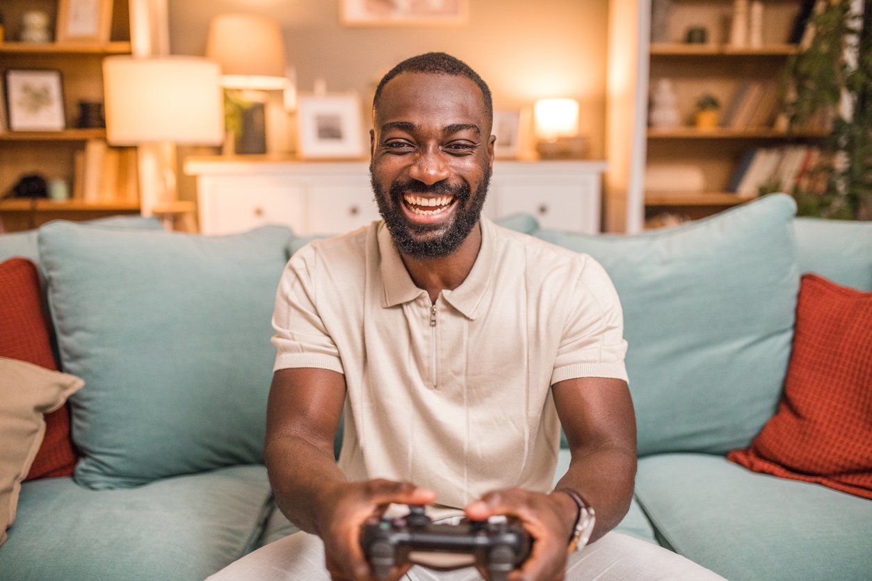 A smiling man sits on a couch holding a video game controller, looking toward the camera. The background shows shelves, framed pictures, and lamps in a cozy living room setting.