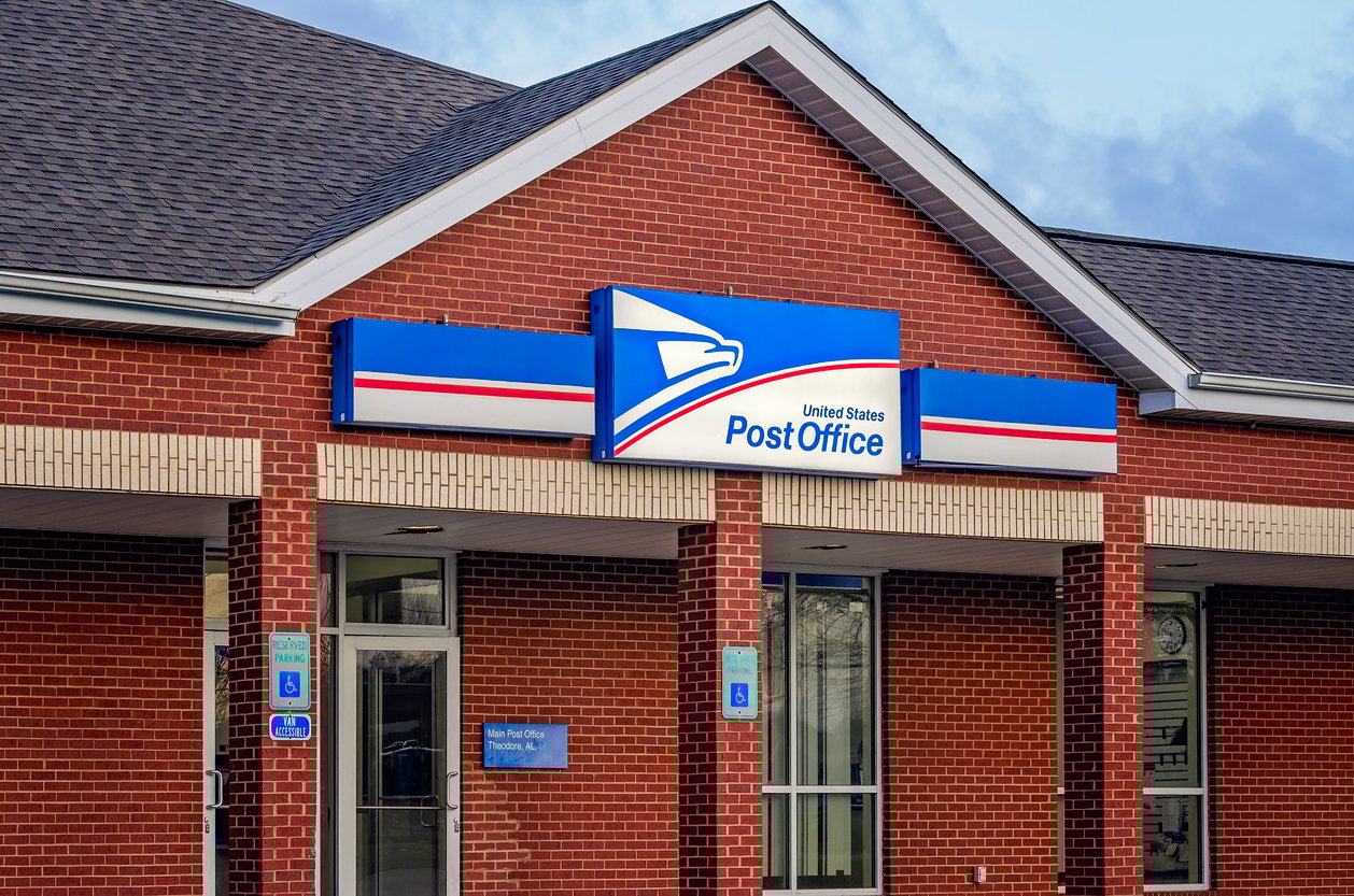 A brick building with a blue and white United States Post Office sign over the entrance, featuring the postal service eagle logo and accessible entrance signs.
