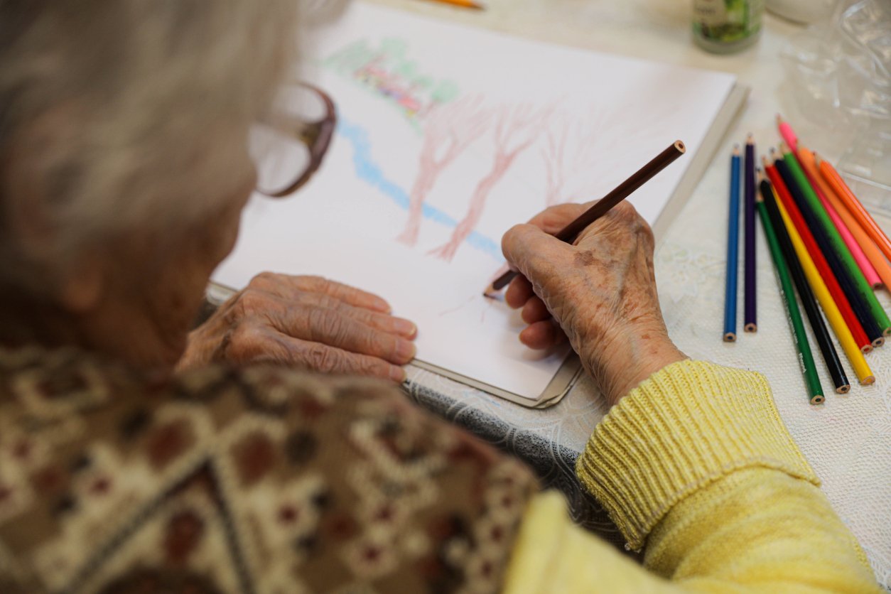 An elderly person wearing glasses and a yellow sweater draws a landscape with trees and a river using colored pencils on white paper; several colored pencils are scattered on the table nearby.