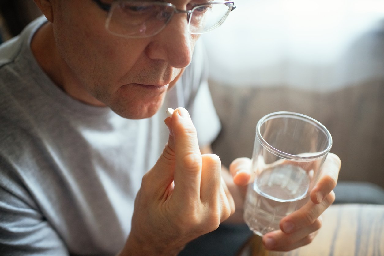 A person wearing glasses holds a pill in one hand and a glass of water in the other, appearing to take medication.