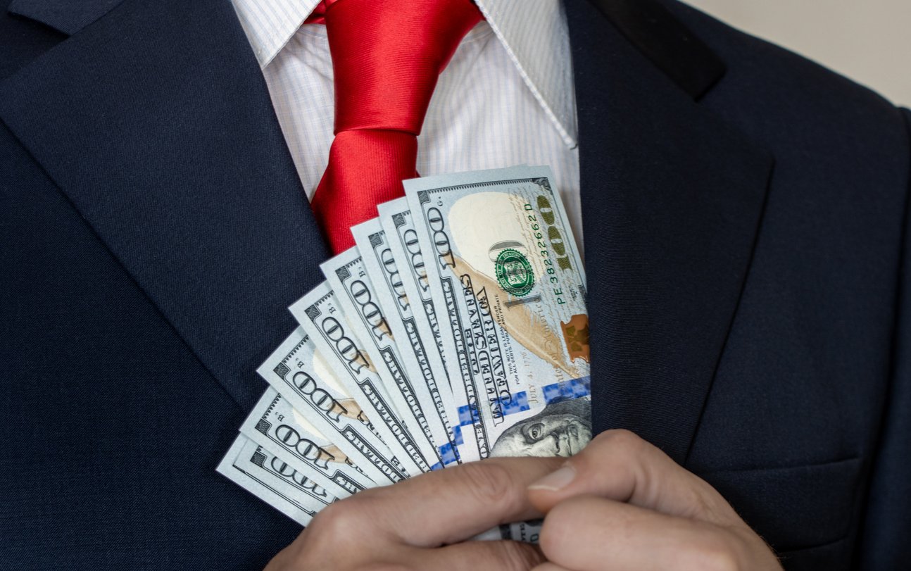 A person in a dark suit and red tie holds a fanned stack of US hundred-dollar bills, partially tucked into the suit jacket.