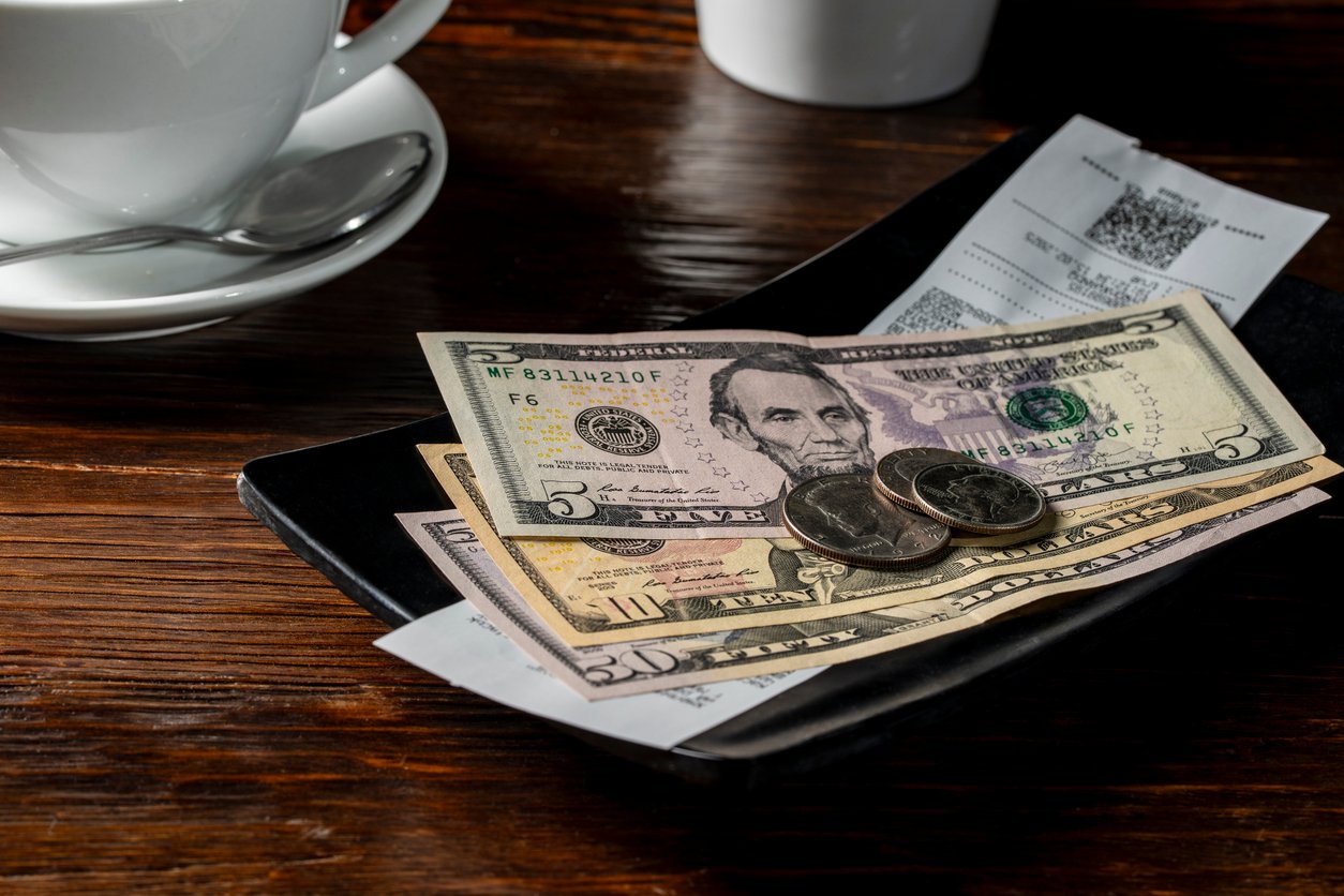 A close-up of a restaurant bill on a black tray with U.S. paper currency, coins, and a receipt. A white coffee cup and saucer are partially visible in the background on a wooden table.