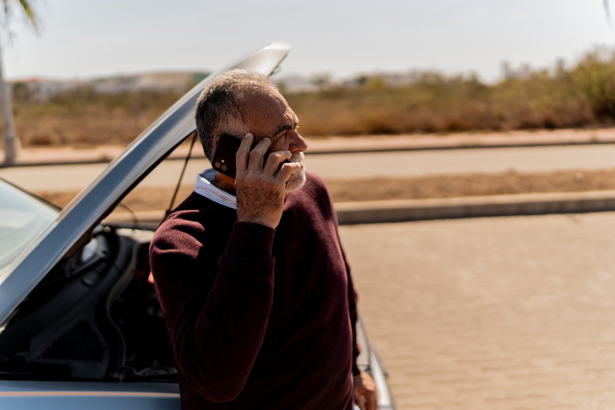 An older man stands next to a car with the hood open, talking on his phone, possibly seeking help. The scene is outdoors on a sunny day with a blurred background of bushes and pavement.