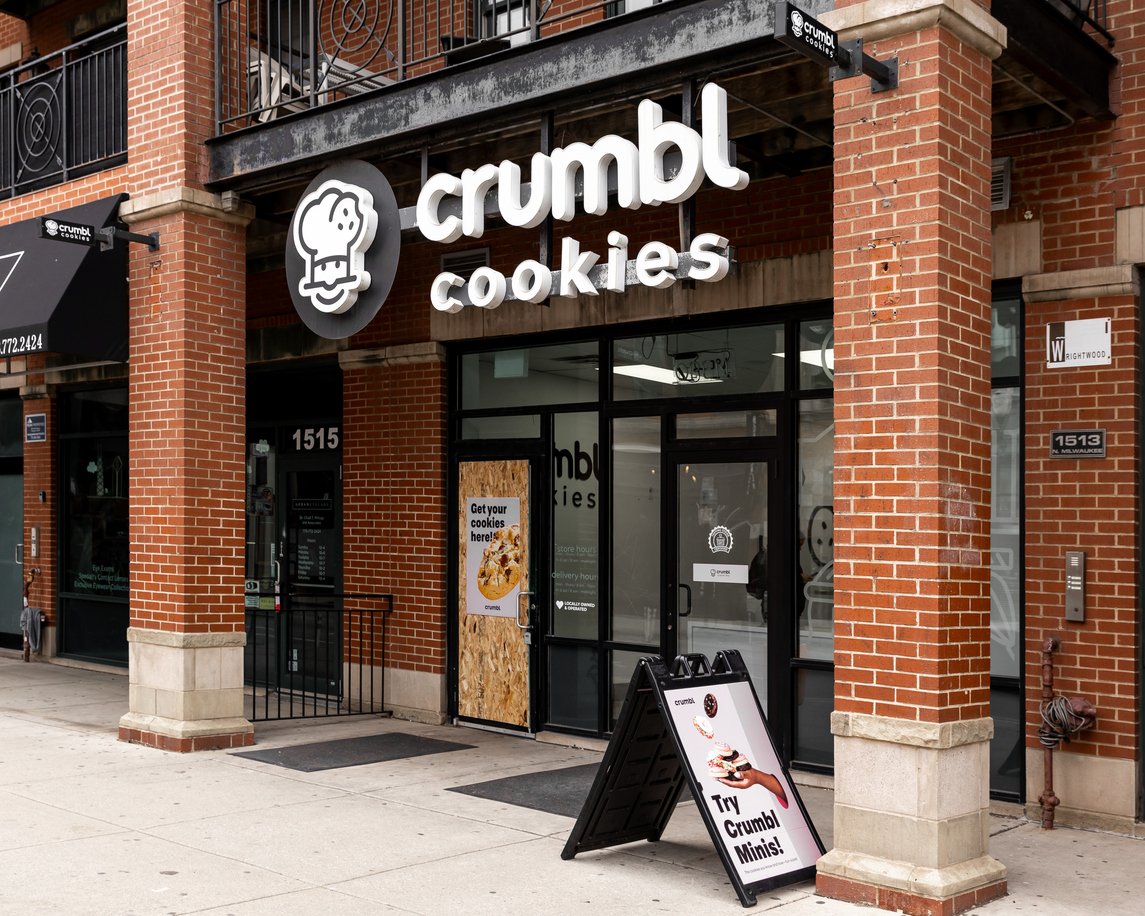 Storefront of Crumbl Cookies with a large sign above the entrance and a sidewalk sign promoting “Crumbl Minis.” The storefront has glass doors and brick exterior. A poster is displayed on one of the front windows.