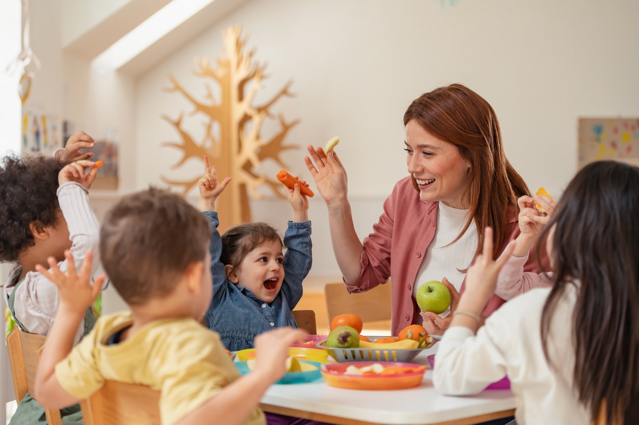 A group of young children and a smiling woman sit around a table holding up pieces of fruit and vegetables, appearing happy and engaged in a bright, cheerful classroom with a wooden tree decoration in the background.
