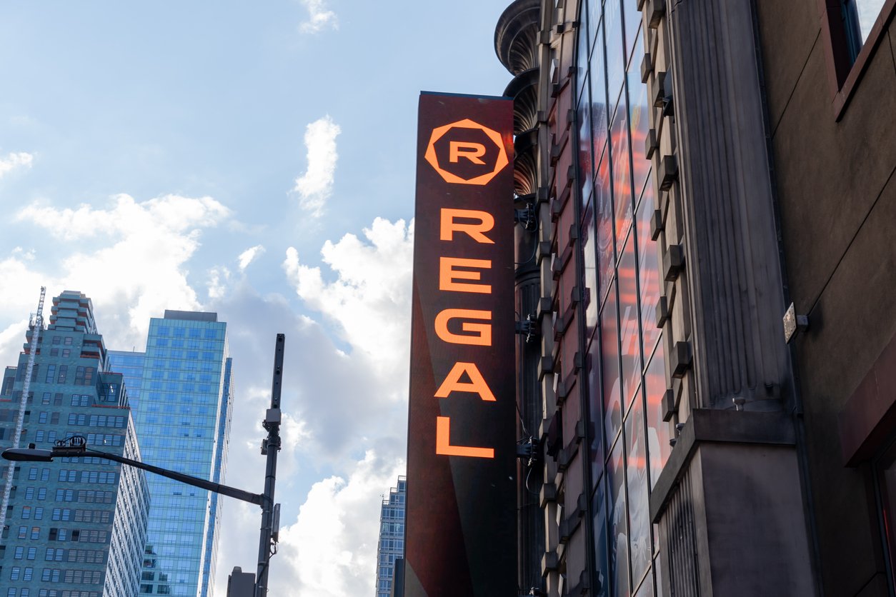 A vertical sign reading "REGAL" in bright orange letters is attached to the side of a city building, with tall skyscrapers and a partly cloudy sky in the background.
