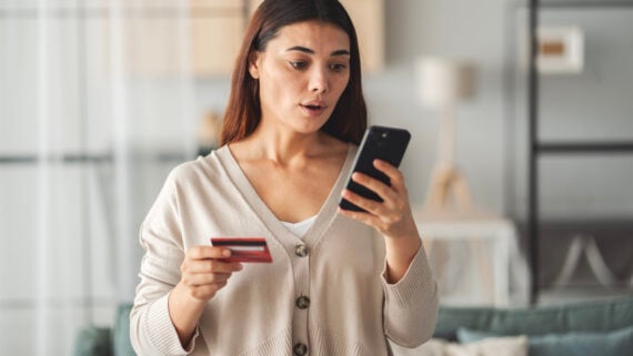 A woman holding a credit card and looking surprised at her smartphone, standing indoors in a living room.