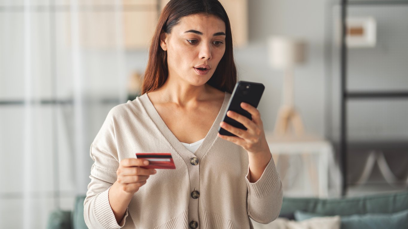 A woman holding a credit card and looking surprised at her smartphone, standing indoors in a living room.