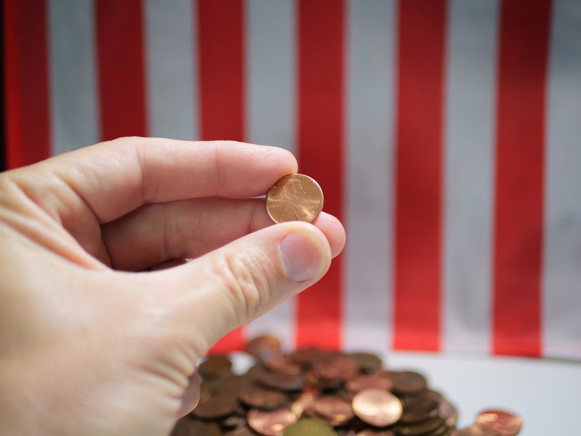 A hand holds a penny in front of a pile of pennies, with a red and white striped background resembling part of the American flag.