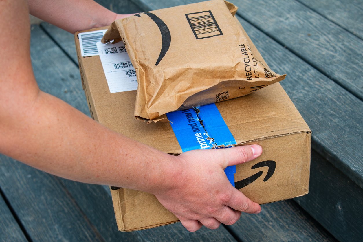 A person holding two Amazon packages, one small and one large, while standing on wooden steps. The packages feature Amazon logos and shipping labels.