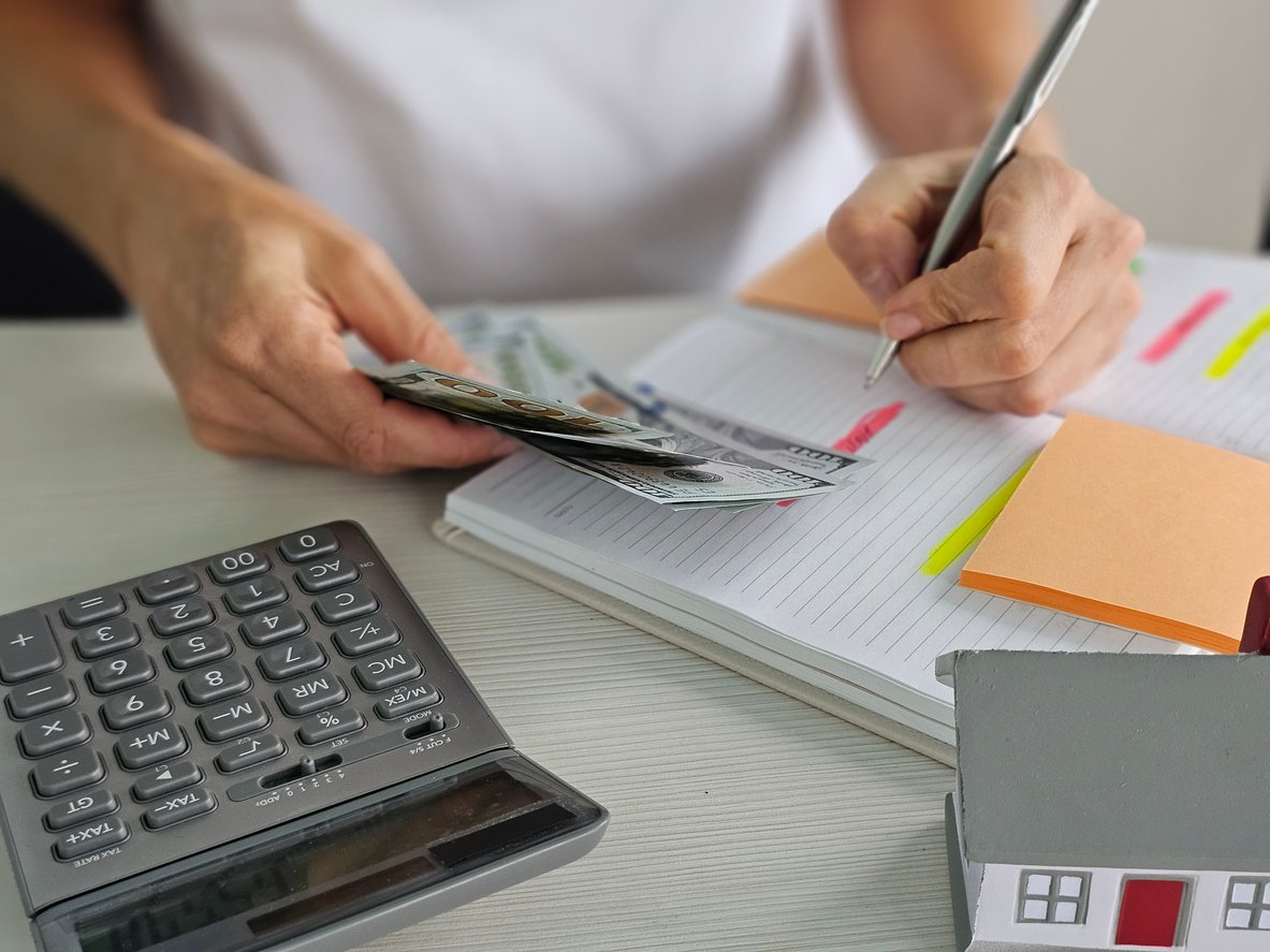 A person counts cash while writing in a notebook. A calculator, sticky notes, and a small model house are on the desk, suggesting budgeting or financial planning activities.
