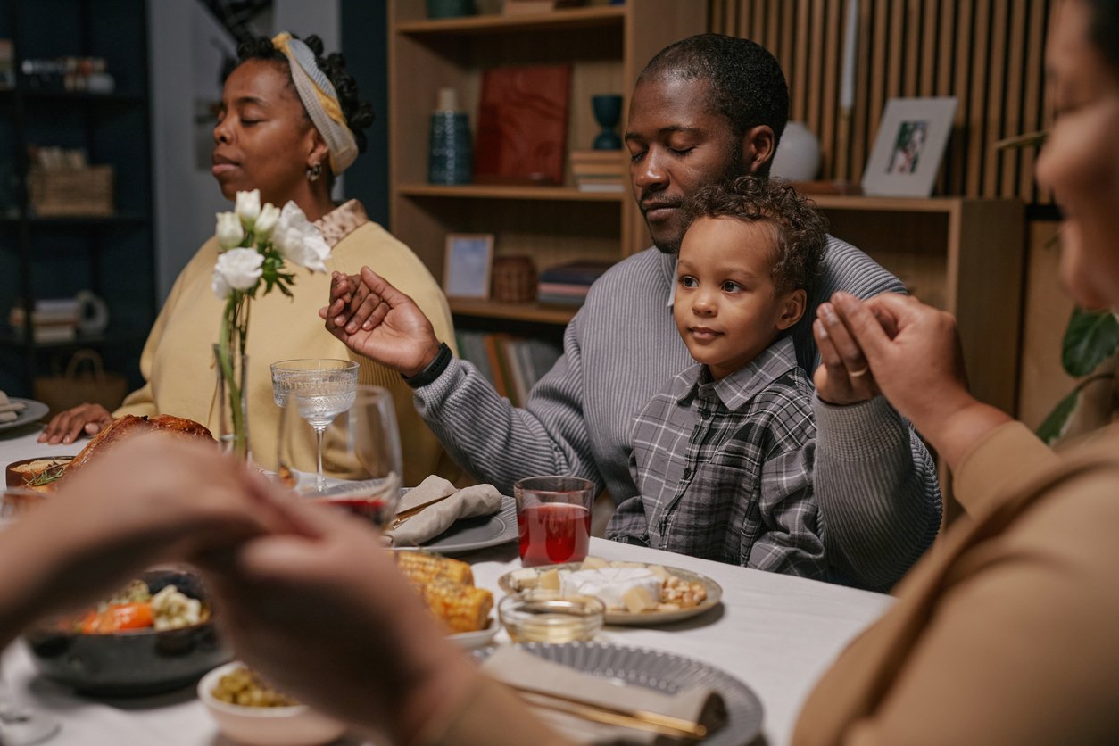 A family sits around a dinner table holding hands in prayer, sharing a peaceful, warm moment before their meal. Their gathering reflects the cherished tradition and changing thanksgiving dinner cost through history.