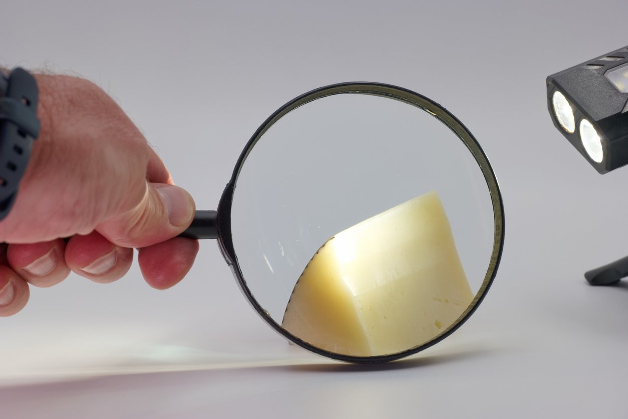 A hand holds a magnifying glass over a wedge of cheese, making the cheese appear larger. A small light with two bulbs illuminates the scene from the right. The background is plain and light-colored.