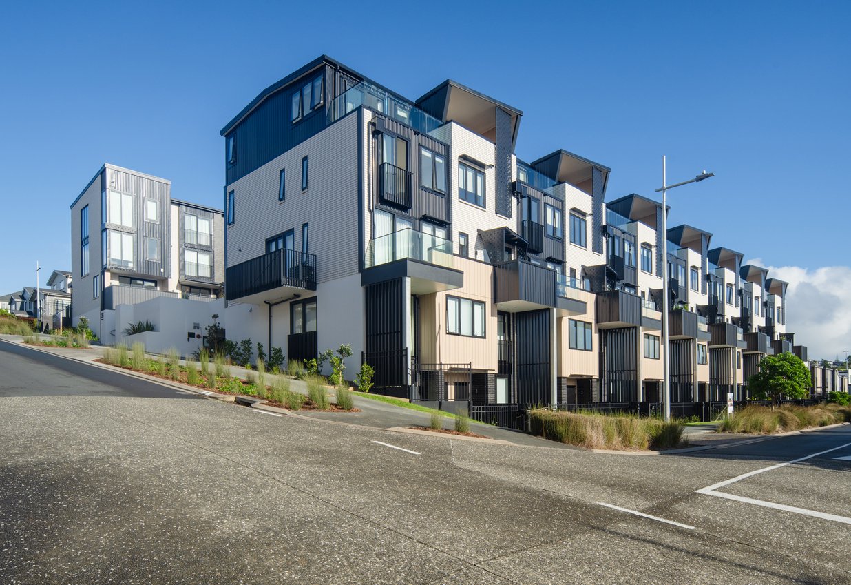 Modern townhouses with white and black exteriors line a sloped street under a clear blue sky, featuring balconies, large windows, and landscaped greenery along the sidewalk.