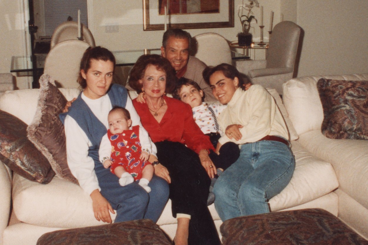 A family of six poses closely together on a white couch in a living room, perhaps after discussing Thanksgiving Dinner Cost History. Two older adults sit in the center, surrounded by three young women and a baby in festive clothing, all creating a warm atmosphere.