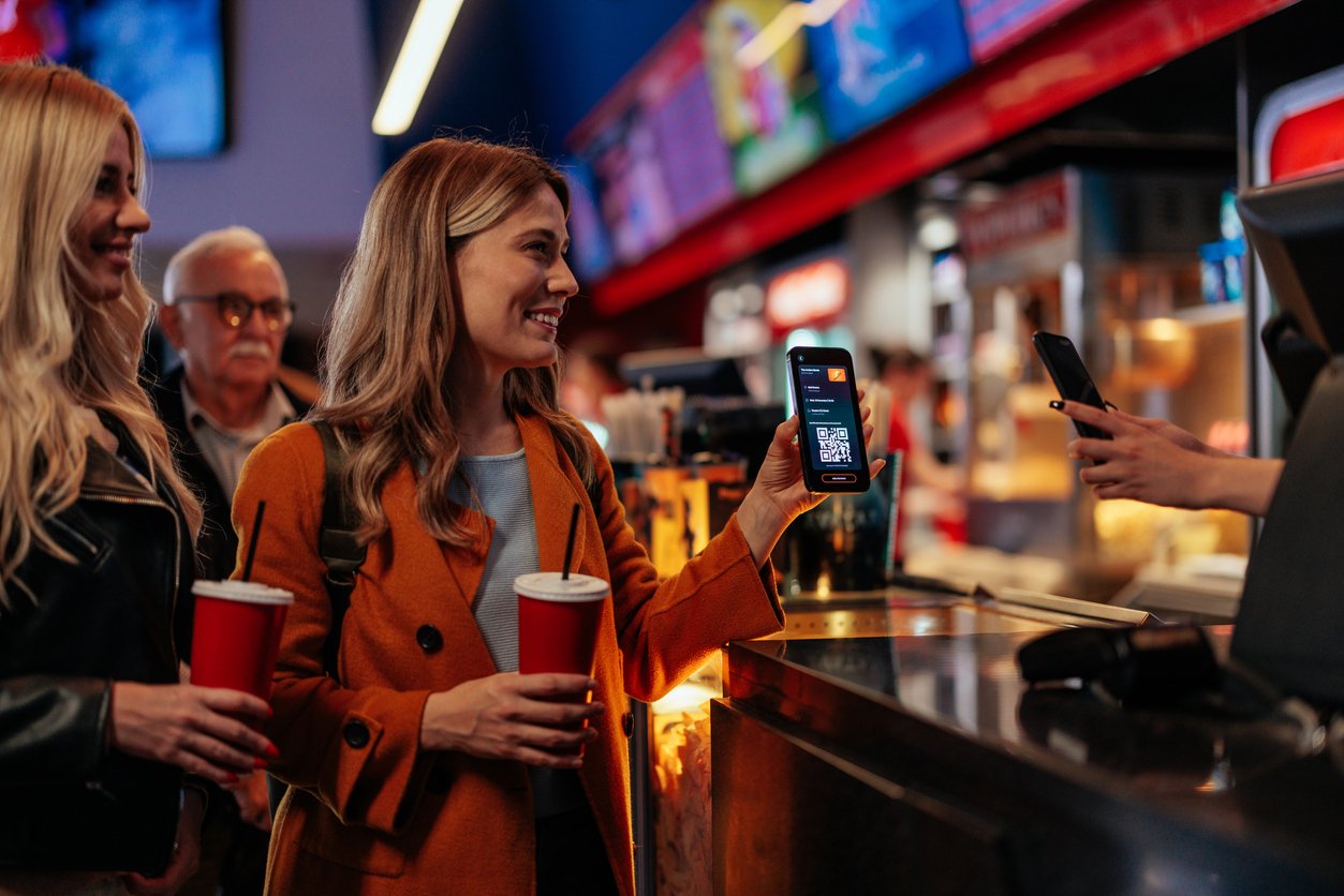 A smiling woman in an orange coat holds a drink and shows her phone with a QR code at a concession stand, while another person scans it. A friend and other people wait in line behind her.