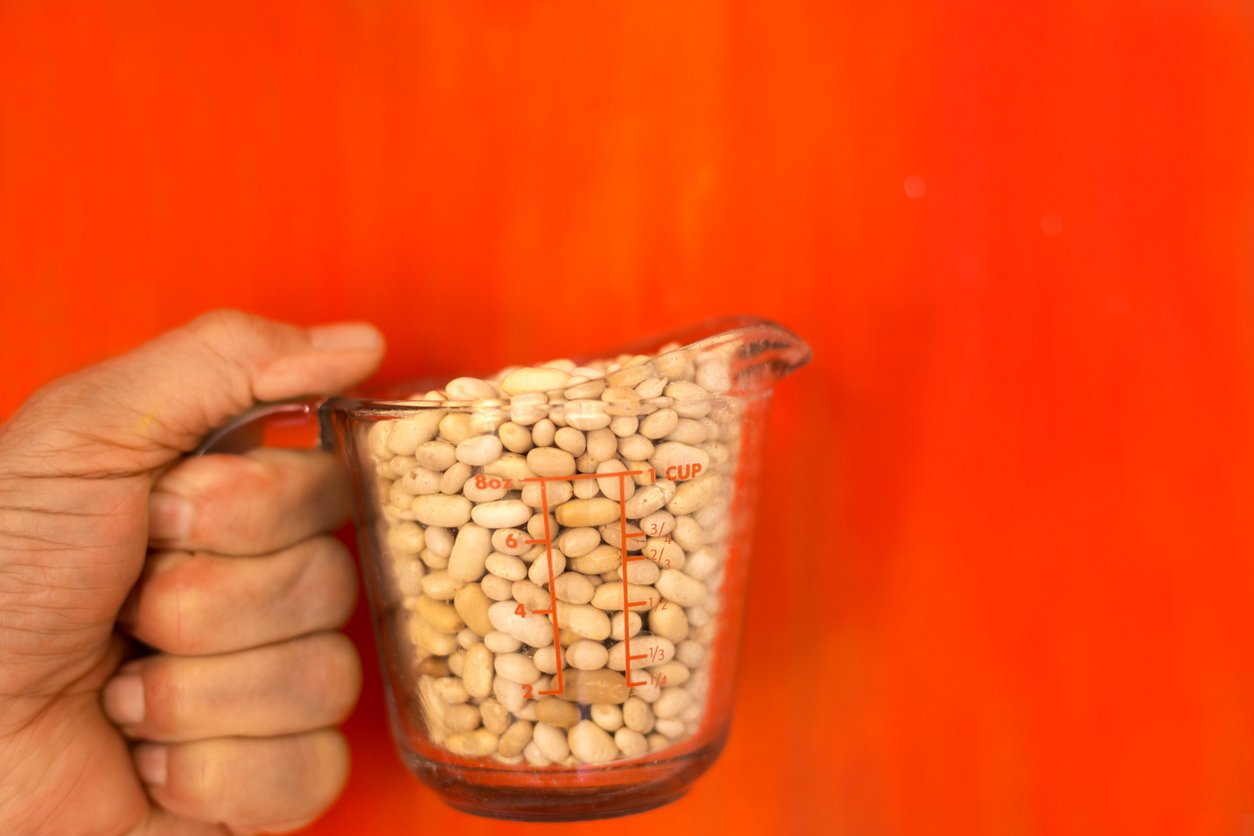 A hand holds a glass measuring cup filled with white beans against a vibrant orange background. The cup shows measurement markings in red.
