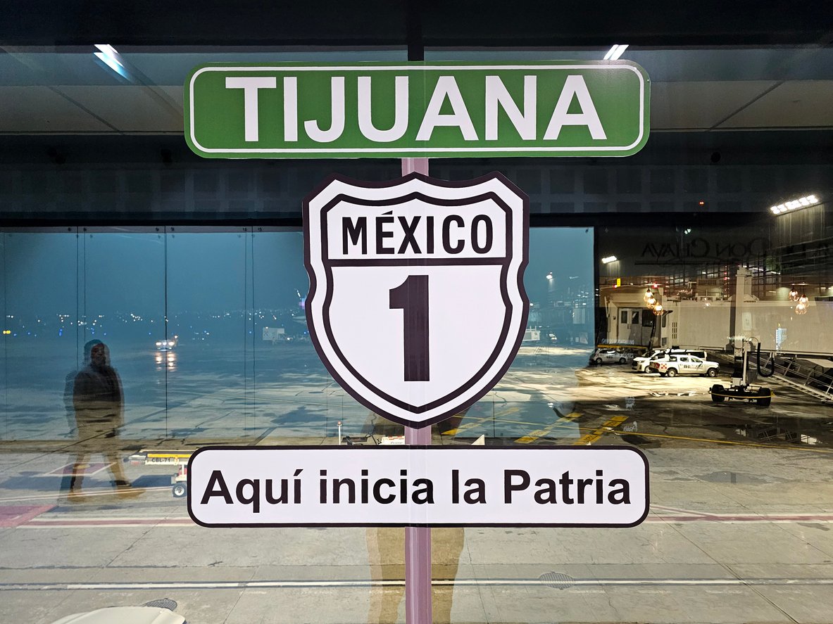 A sign at an airport window reads "TIJUANA," "México 1," and "Aquí inicia la Patria" ("Here the homeland begins"), with the runway and airport vehicles visible outside.