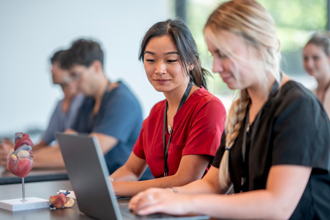 Two medical students sit at a desk, one in red scrubs and one in black, working on a laptop in a classroom setting. Other students and a heart model are visible in the background.