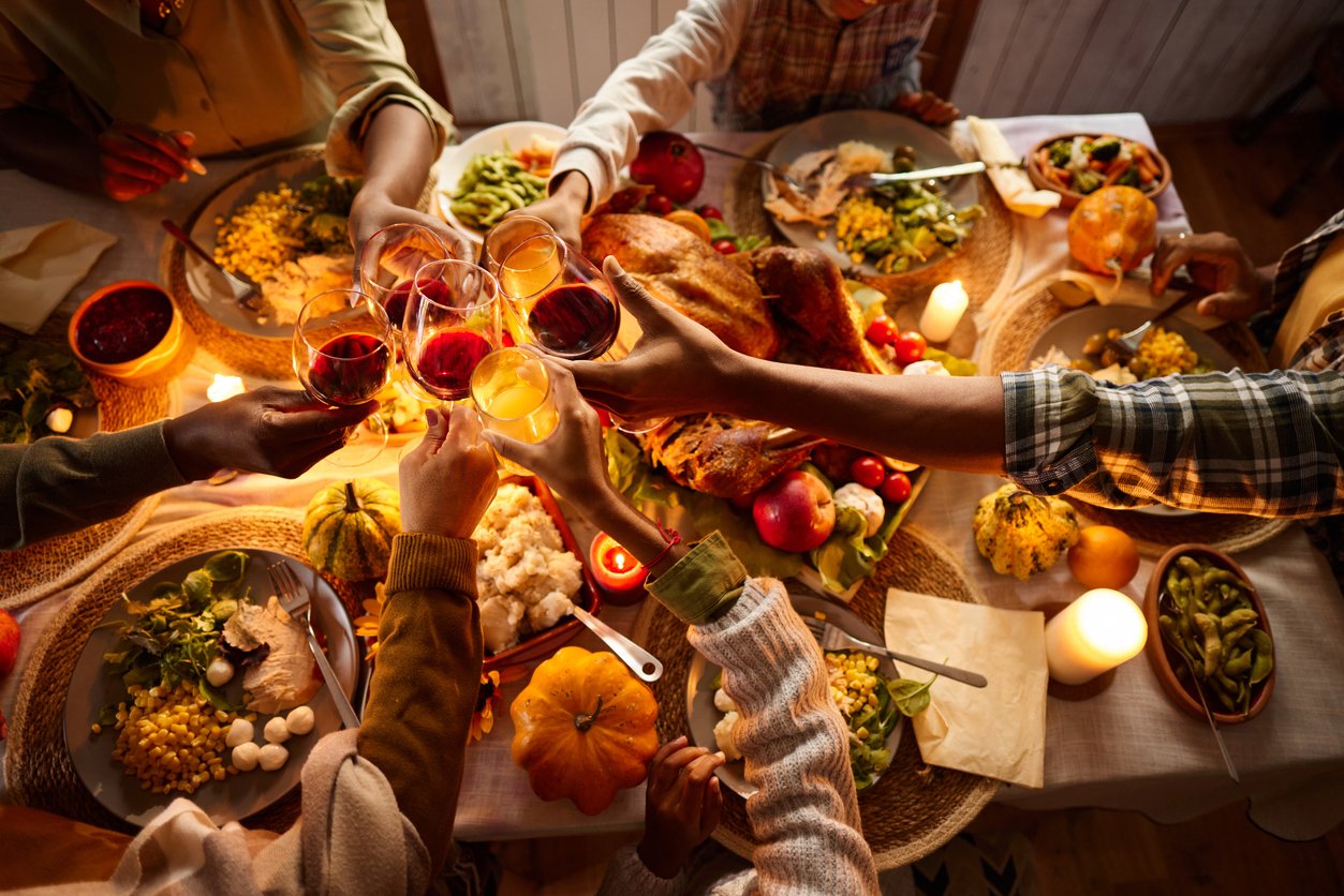 A group of people sitting around a festive table filled with Thanksgiving food, raising their glasses for a toast. The table is decorated with candles, pumpkins, and a roasted turkey.
