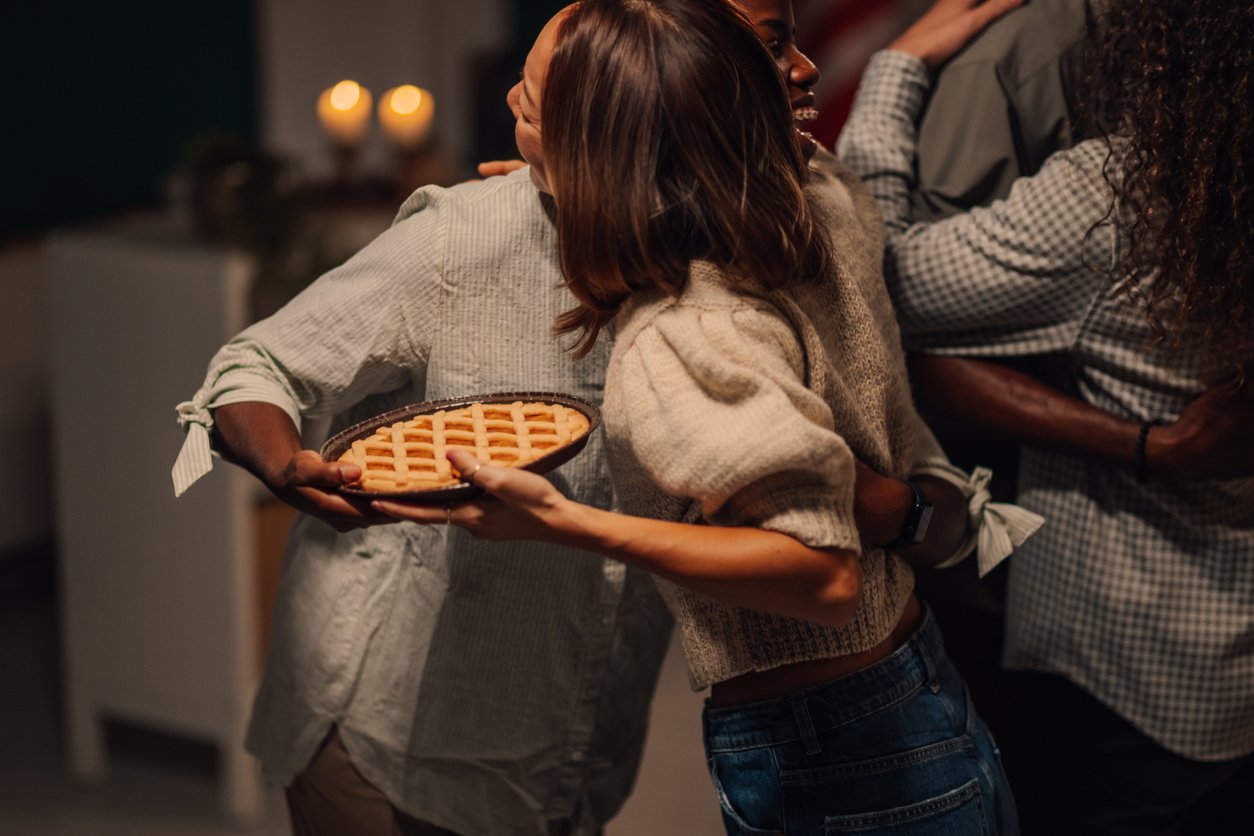 Two people hug warmly while one holds a freshly baked lattice pie, capturing the joy of gathering despite rising Thanksgiving dinner cost. Two others join in, creating a cozy, celebratory atmosphere with soft lighting in the background.