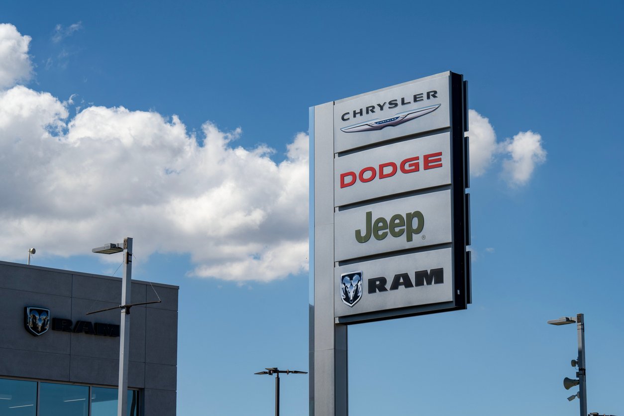 A tall outdoor sign displays the logos of Chrysler, Dodge, Jeep, and Ram against a blue sky with clouds, next to a dealership building.