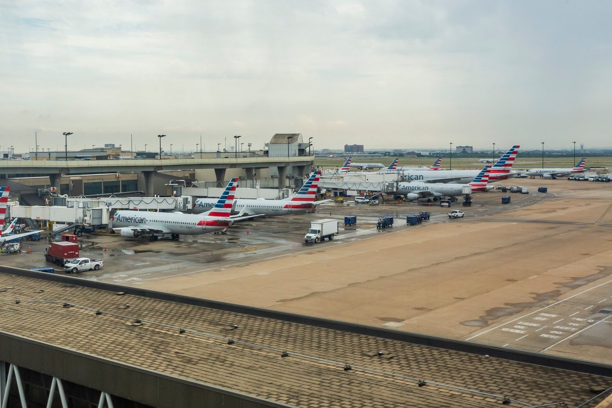 Multiple American Airlines airplanes are parked at gates at an airport terminal, with ground vehicles and equipment nearby on a cloudy day. The scene overlooks the runway and surrounding airport infrastructure.