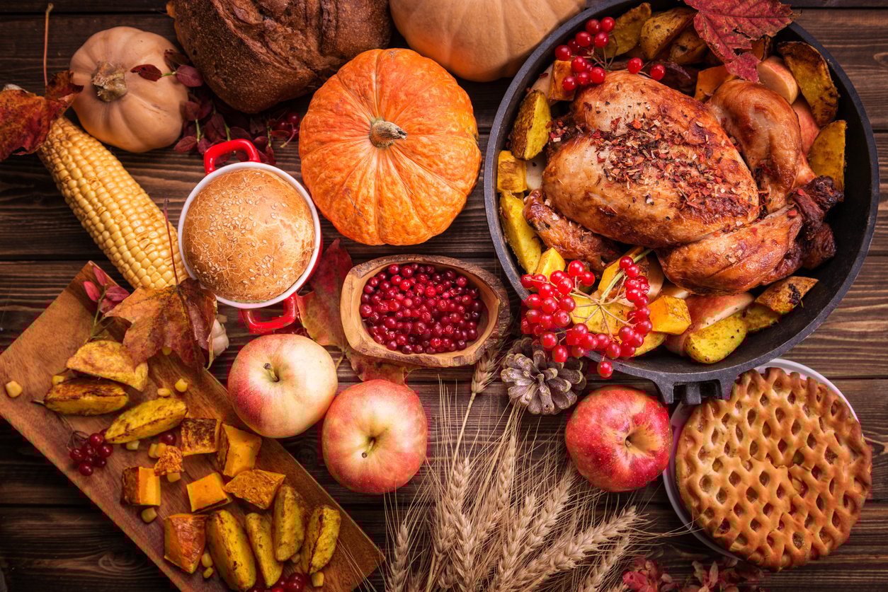 A festive autumn meal inspired by Thanksgiving dinner cost history, featuring roasted chicken, baked vegetables, apples, corn, pumpkins, cranberries, pie, bread, and autumn leaves arranged on a wooden table.