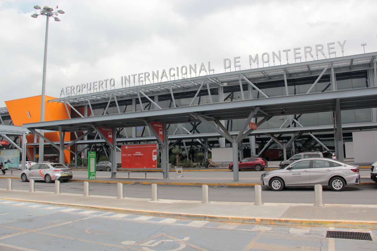 Exterior view of Monterrey International Airport, showing its modern facade, parked cars in front, and a sign reading "Aeropuerto Internacional de Monterrey" on the roof.