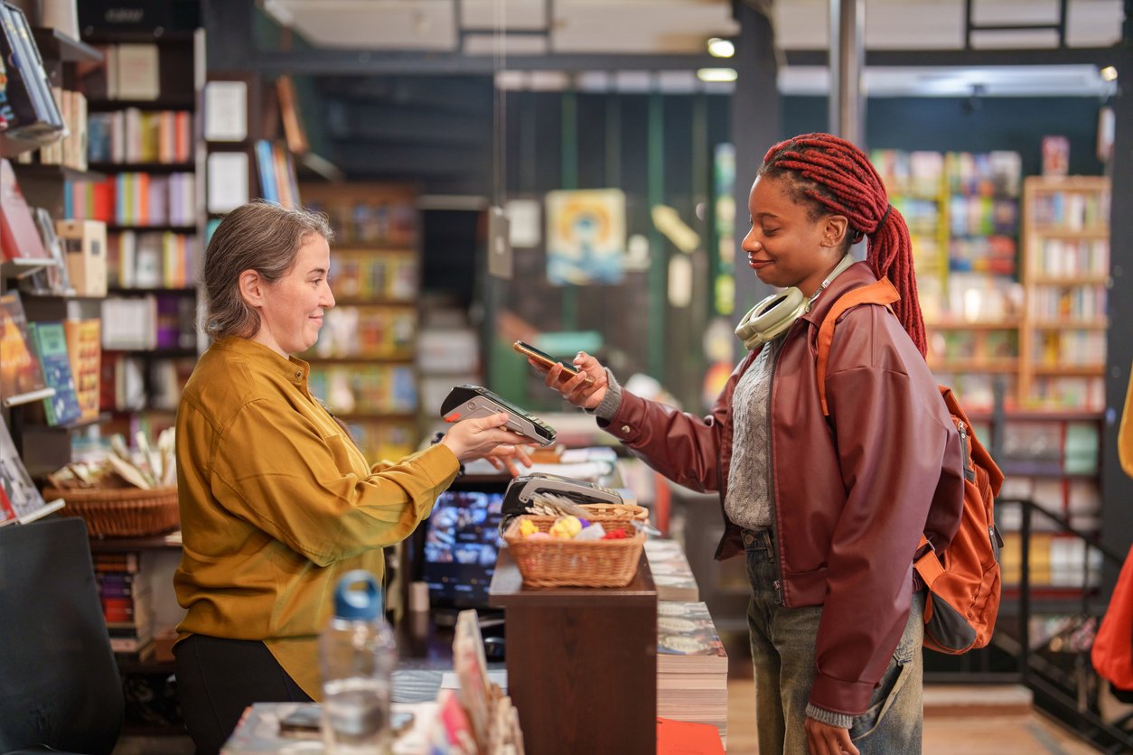 A young woman with headphones pays with her smartphone at a bookstore counter, smiling at the cashier who is holding a card reader. Shelves of books fill the background.