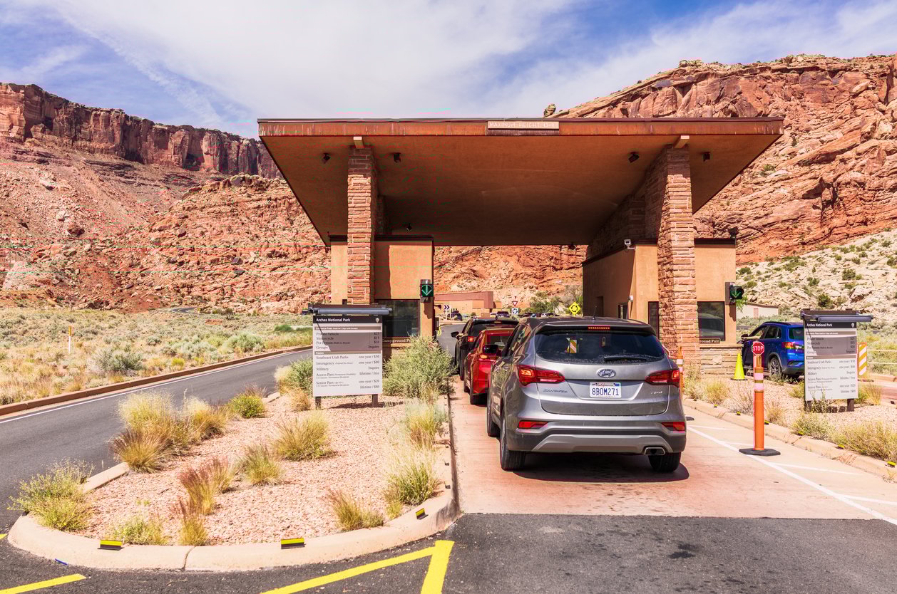 Cars wait in line at a national park entrance booth in a desert landscape with rocky red cliffs in the background. Signs and vegetation are visible along the paved road.